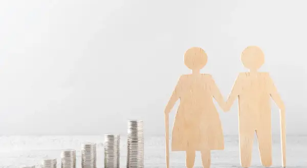 Drewniane figurki starszej pary z laskami, obok rosnących stosów monet. Symbolizuje to finansowe wsparcie i opiekę na członka rodziny w podeszłym wieku.