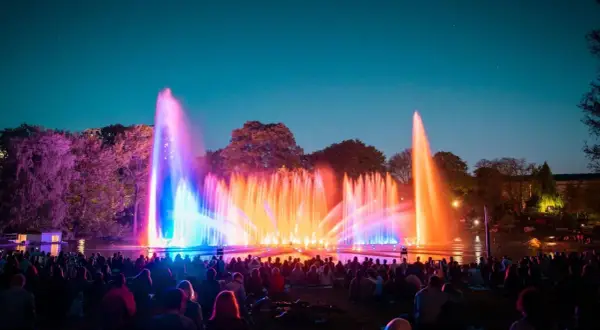 Spektakuläre Wasserspiele in Planten un Blomen erhellen den Abendhimmel in bunten Farben. Eine Menschenmenge sitzt im Gras und bestaunt die Lichtshow.