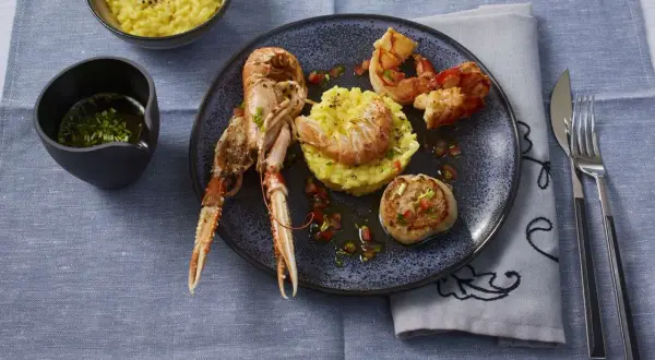 Ein Teller mit frutti di mare: Kaisergranat, Garnelen und Jakobsmuschel auf Safranrisotto, garniert mit Tomatenwürfeln. Daneben ein Schälchen mit Sauce und ein Schälchen Risotto.