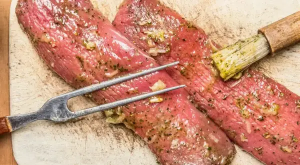 Polędwiczki wieprzowe w aromatycznej marynacie, gotowe do pieczenia. Widelec i pędzelek do sosu czekają.