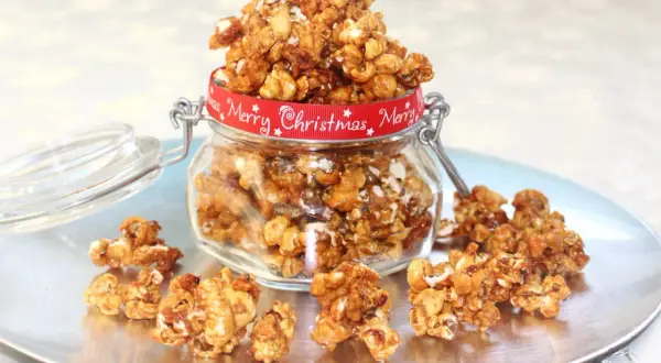 En burk fylld med karamelliserade popcorn, dekorerad med ett rött band med texten "Merry Christmas". Några popcorn ligger utspridda på ett fat.