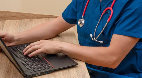 Lekarz w niebieskim fartuchu z czerwonym stetoskopem pisze na laptopie, szukając informacji, jaki lekarz zarabia najwięcej.