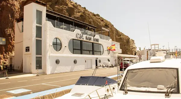 Edificio blanco con el letrero "C.N. SAN JOSE" y una bandera española ondeando. Un restaurante con vistas al mar, ideal para disfrutar de la brisa marina.