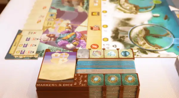 Spielkomponenten für das Dead Reckoning Brettspiel, darunter Karten, Marker und Würfel, sind ordentlich in braunen Halterungen gestapelt.