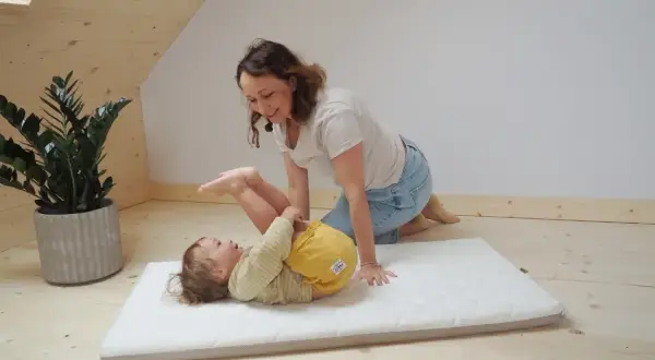 Mama bawi się z dzieckiem na miękkim materacyku do łóżeczka. Radosny maluch leży na plecach, a mama klęczy obok, uśmiechając się.