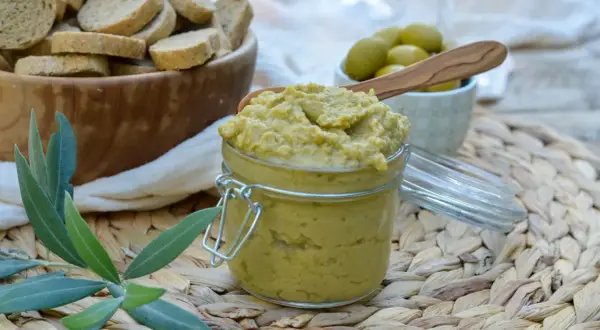 Un pot de tapenade verte, garni d'une cuillère en bois, est prêt à être dégusté avec des toasts et des olives.
