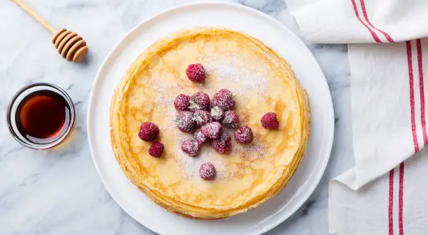 Ein Stapel goldbrauner Ofenpfannkuchen, garniert mit Himbeeren und Puderzucker. Daneben eine Schale Honig und ein Honiglöffel. Perfekt für den Thermomix!