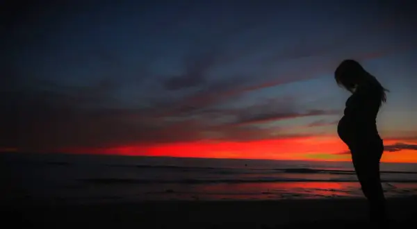 Kobieta w ciąży na tle zachodu słońca nad oceanem. Pamiętaj o ubezpieczeniu turystycznym w ciąży, by podróżować bezpiecznie.