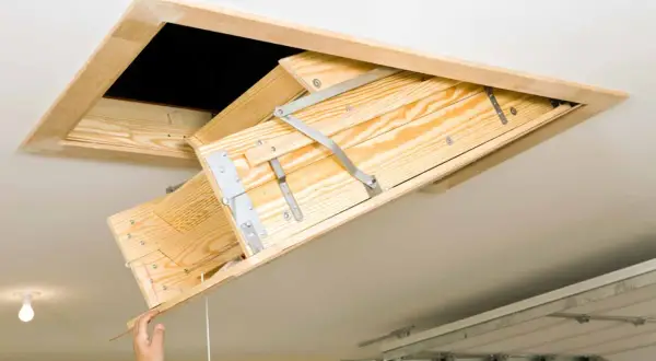 A wooden attic ladder is being installed, with a hand pulling down the first section.