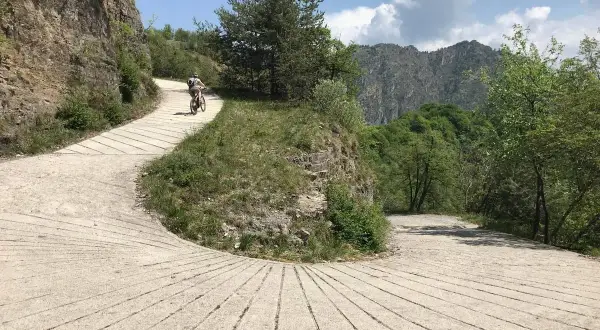 Ciclista su strada sterrata nel Parco Alto Garda Bresciano, tra rocce e vegetazione lussureggiante, con montagne sullo sfondo.