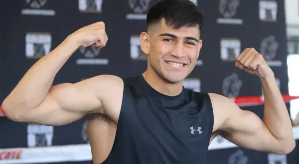 Alexis boxer flexes his biceps, smiling confidently in a black tank top.