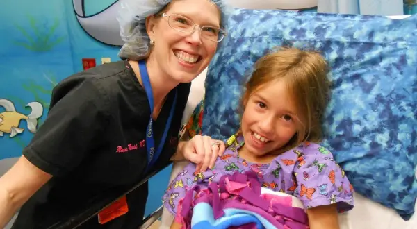 Niña sonriente en cama de hospital, junto a enfermera. Ella tiene síndrome de Marfan y recibe cuidados.