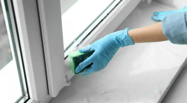 Ręka w niebieskiej rękawiczce czyści parapet gąbką, pokazując, czym usunąć klej po taśmie klejącej.