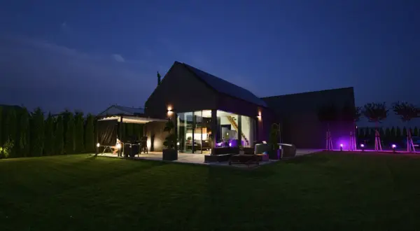 Nowoczesny dom pasywny oświetlony nocą. Widać taras z grillem, duże okna, ogród i fioletowe lampy.