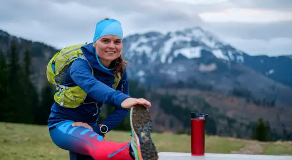 Uśmiechnięta biegaczka przygotowuje się do treningu ultramaraton 50 km, rozciągając nogę na tle gór.