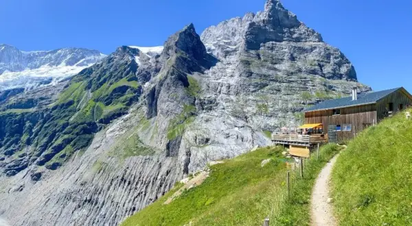 Ein Weg führt zu einer Berghütte mit Terrasse vor imposanter Schweizer Bergkulisse. Perfekt für eine Pause beim **Schweiz wandern**.