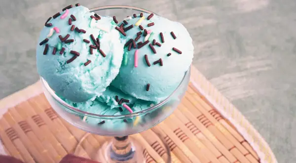 Zwei Kugeln blaues Eis mit bunten Streuseln in einem Glas auf einem Holzuntersetzer.