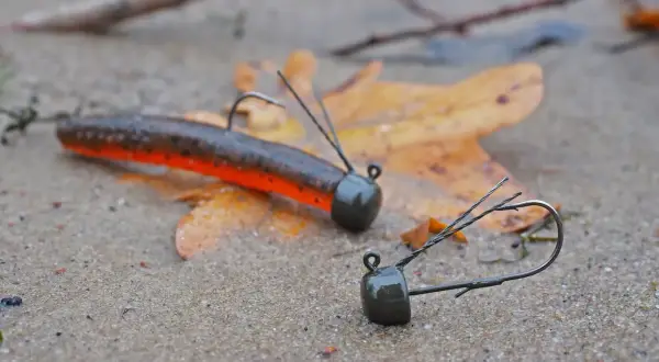 Zwei Angelköder, ein ned rig und ein weiterer, liegen auf nassem Sand neben einem Herbstblatt.