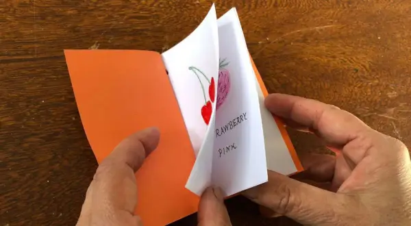 Mani che sfogliano un piccolo libro fatto a mano, con disegni di fragole e ciliegie. Un'idea per imparare come fare un libro a mano.
