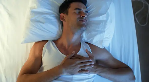 Mann liegt im Bett, hält sich die Brust. Er leidet unter Herzrasen nachts, die Augen sind geschlossen, das Gesicht verzogen.
