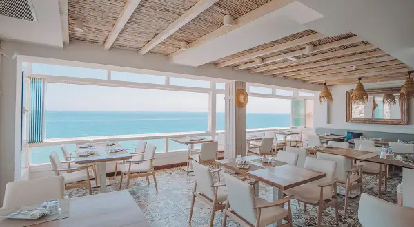 Comedor de la casa Pepe San José con vistas al mar. Mesas puestas, sillas blancas y techo de caña.