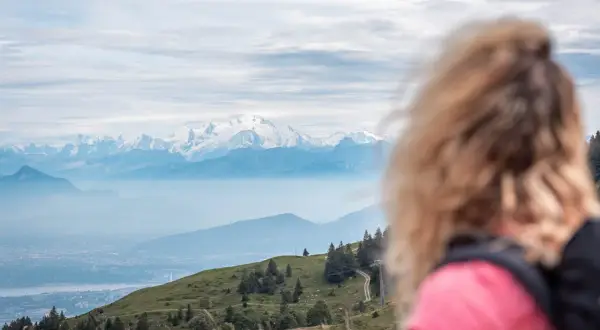 Vue panoramique sur les Alpes enneigées, avec une femme blonde en premier plan. Un sentier serpente sur la colline verdoyante, invitant au trek dans le Jura.
