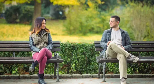 En kvinna och en man sitter på varsin parkbänk och ler mot varandra. Kanske funderar hon på hur blir man attraktiv som tjej, och han på hur blir man attraktiv som kille.