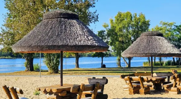 Drewniane stoły z ławami pod strzechą na plaży w Rogoźno Wielkopolskie, nad błękitną wodą.