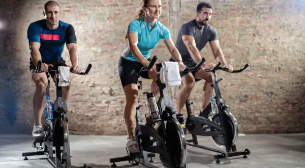 Trzy osoby ćwiczą na rowerach stacjonarnych. To jest właśnie spinning – intensywny trening kolarski w grupie.