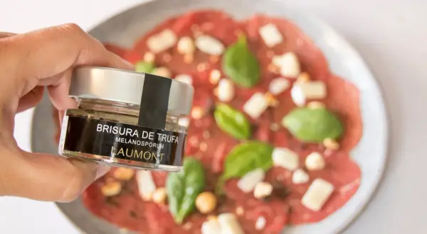 Mano sostiene un frasco de "Brisura de Trufa" Laumont, listo para añadir a un carpaccio de ternera con queso y albahaca.