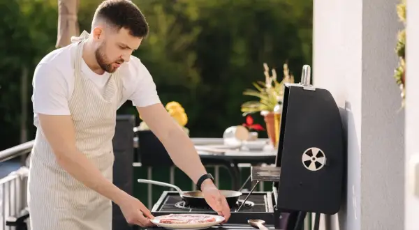 Mężczyzna w fartuchu przygotowuje mięso na grillu na balkonie. Czy można robić grilla na balkonie? Tak, jeśli jest to bezpieczne i zgodne z przepisami.