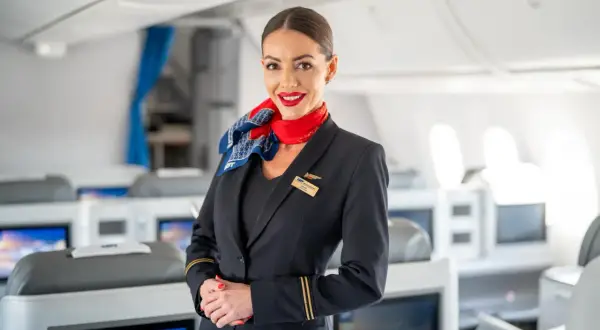 Uśmiechnięta członkini **cabin crew** w granatowym mundurze z czerwonym szalem, stoi na tle rzędów foteli w samolocie.