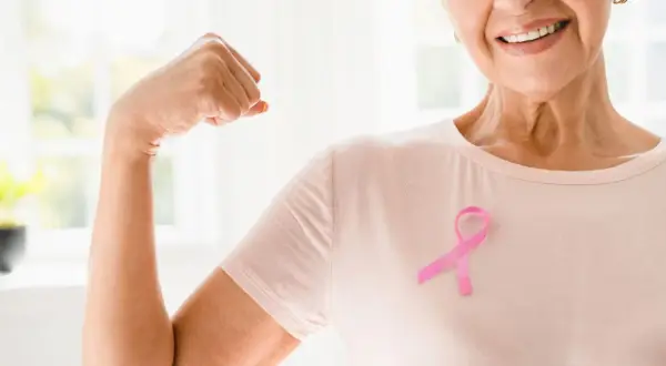 Lächelnde Frau mit rosa Schleife zeigt Bizeps. Onkologische Physiotherapie stärkt den Körper und gibt Hoffnung.