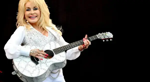 Dolly Parton, w białym, błyszczącym stroju, gra na gitarze. Wyobraź sobie, że śpiewa "Jolene", a tekst piosenki rozbrzmiewa w powietrzu.