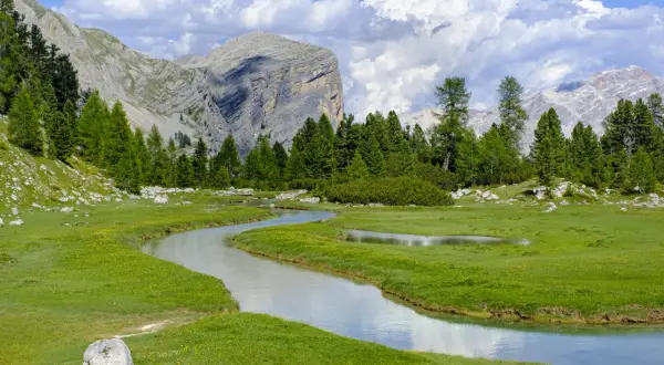 Un ruscello serpeggia tra prati verdi e fiori gialli, con imponenti montagne sullo sfondo. Un paesaggio da cartolina, tipico delle località di montagna famose.