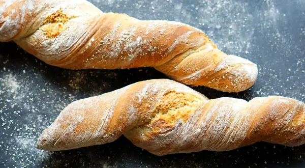 Zwei gedrehte Brote, die an ein Wurzelbrot Rezept dunkel erinnern, liegen auf dunklem Untergrund, bestäubt mit Mehl.
