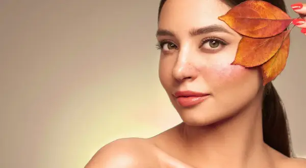 Une femme au teint légèrement rougi, cachant une partie de sa joue avec des feuilles d'automne. Une astuce pour savoir comment enlever des rougeurs rapidement.