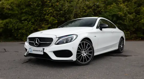 Biały Mercedes C43 AMG coupe z czarnymi felgami, zaparkowany na tle zieleni.