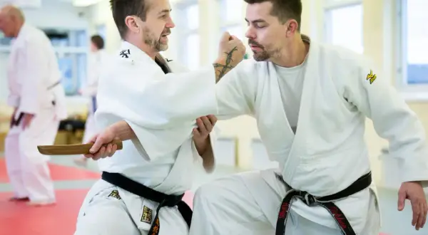 Två män i judodräkter tränar en judo kast. En av dem håller i ett träföremål.