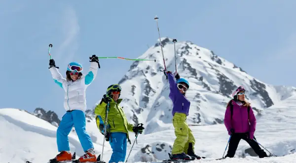 Famille en tenue de ski, bras levés, profitant du sport alpin sur une pente enneigée avec des montagnes majestueuses en arrière-plan.
