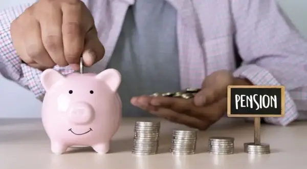 Oszczędzanie na emeryturę pomostową: ręka wrzuca monetę do świnki skarbonki, obok stosy monet i tabliczka z napisem "PENSION".