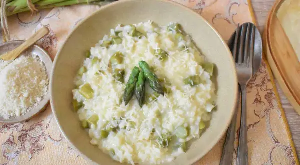 Kremowe risotto ze szparagami, posypane parmezanem, z kilkoma świeżymi szparagami na wierzchu. Obok miseczka z tartym serem.
