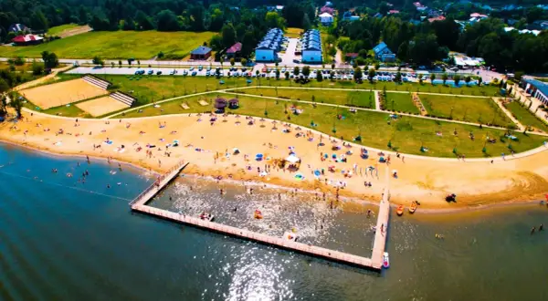 Słoneczny dzień nad zalewem żyrardowskim. Ludzie odpoczywają na plaży, kąpią się w wodzie i spacerują po molo.