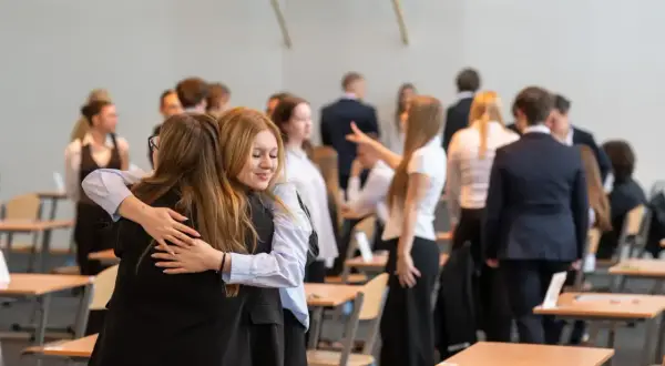 Dwie uczennice przytulają się, gdy wyniki matur są ogłaszane. W tle widać innych uczniów i sale lekcyjne.