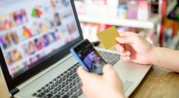 Dłoń trzymająca kartę płatniczą i telefon nad klawiaturą laptopa.