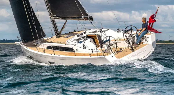 Un uomo guida una barca a vela bianca con vele nere, solcando le onde. Il nome "Xelebration" è visibile sullo scafo.