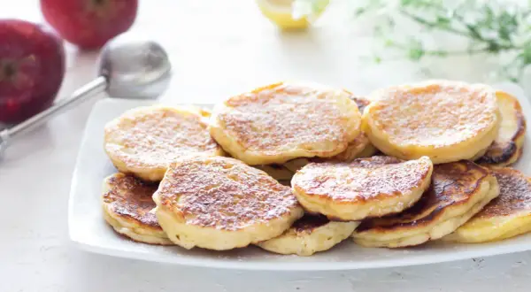 Piatto di frittelle di mele dorate e soffici, spolverate di zucchero a velo. Sullo sfondo, mele rosse e un colino.