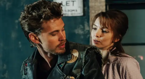 Ein Mann mit Lederjacke und eine Frau im rosa Hemd blicken sich an. Szene aus "The Bikeriders" Stream.