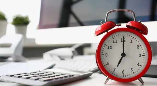 Sulla scrivania, un orologio rosso segna le 7. La calcolatrice e il computer suggeriscono che il giorno di riposo viene pagato, ma il lavoro attende.