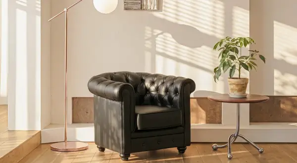 En svart chesterfieldfåtölj står bredvid ett runt sidobord med en grön växt. En kopparfärgad golvlampa ger ett mjukt ljus.